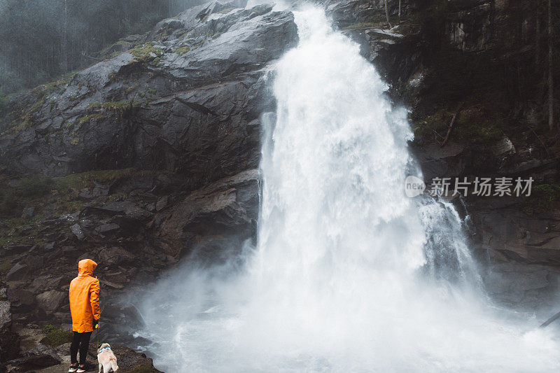 一个穿着雨衣的女人和一只狗在奥地利阿尔卑斯山观看巨大的克里姆尔瀑布