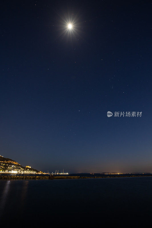 长曝光的夜晚拍摄的天空在平静的海面上与城镇的灯光，明亮的月亮和星星