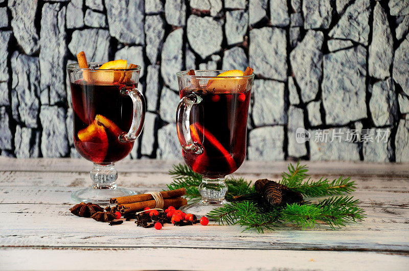 这是一款温馨的圣诞礼物，桌上放着两杯加了香料的热红酒，周围是冷杉枝、松果、丁香、肉桂枝和红浆果。