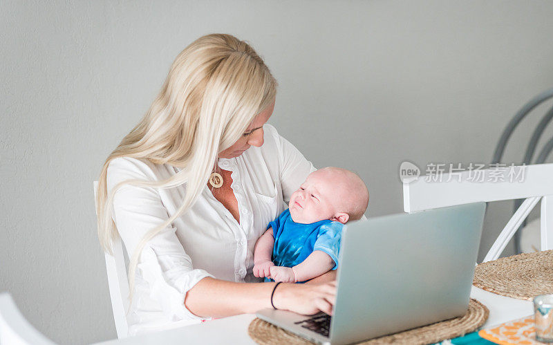 一名妇女抱着哭闹的婴儿在家里用笔记本电脑工作。在养育子女和工作与生活之间平衡的母亲