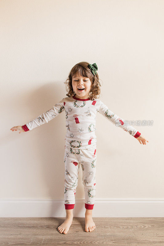 2022年12月，一个快乐而充满希望的三岁小女孩闭着眼睛微笑，穿着简单的中性圣诞睡衣和深绿色发结，在家里度过一个舒适的圣诞节