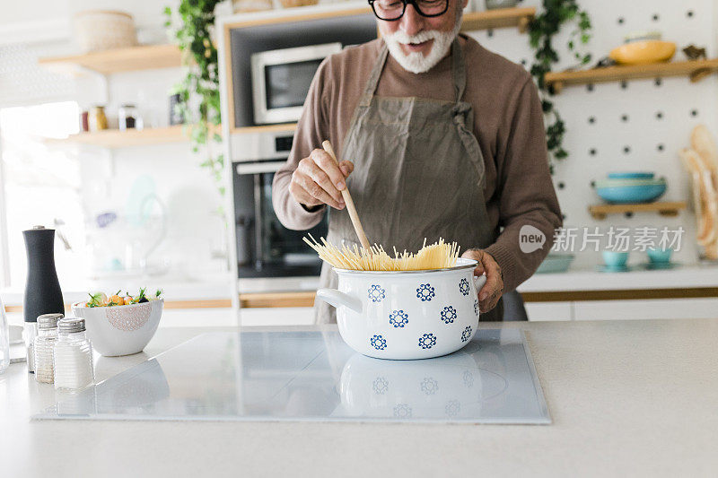 一个快乐的老人站在厨房柜台前，一边用勺子搅拌沙拉，一边在炉子上做美味健康的饭菜。