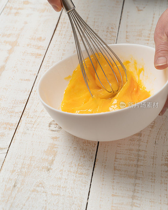 特写:一只手用打蛋器在白色厚木板桌上打鸡蛋