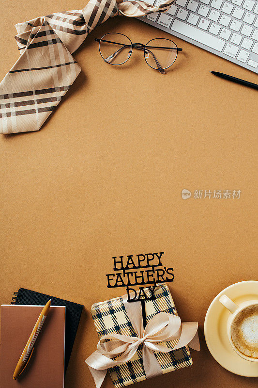 祝父亲节快乐，将礼物盒、领带、纸质笔记本、咖啡杯、键盘、眼镜平铺在棕色的桌面上。