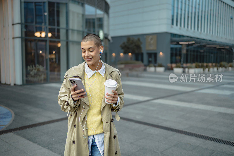 拿着智能手机和咖啡杯的年轻女子在户外环绕着现代建筑