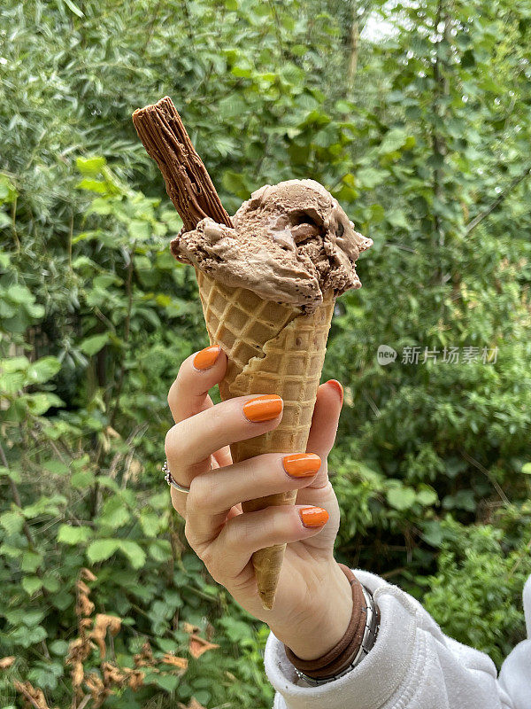 特写照片中，一个无法辨认的女人涂着橙色指甲油，手里拿着一勺巧克力冰淇淋，里面有巧克力片，正在融化的冰淇淋，背景是林地，前景是焦点