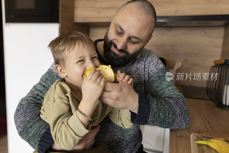 父亲和孩子在厨房分享食物。温暖的联系，美丽的自然光线下充满希望的情感。吃他们最喜欢的水果苹果和橘子。健康的生活方式，亲密的时间。