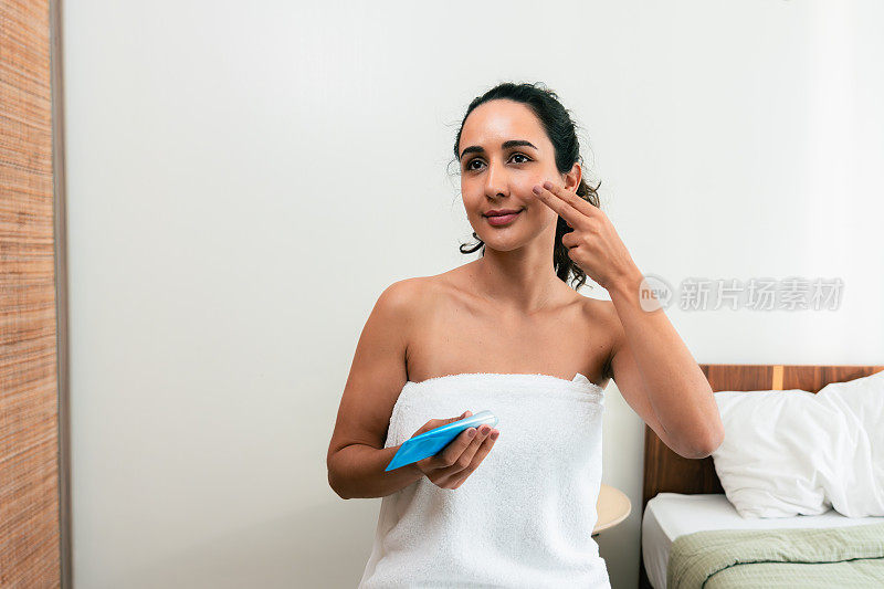 一名微笑的拉丁美洲妇女披着白色毛巾，在家里用她的闲暇时间做身体护理，特别是她的脸，用防晒霜防止晒伤。护肤习惯。使皮肤柔软健康的化妆品。
