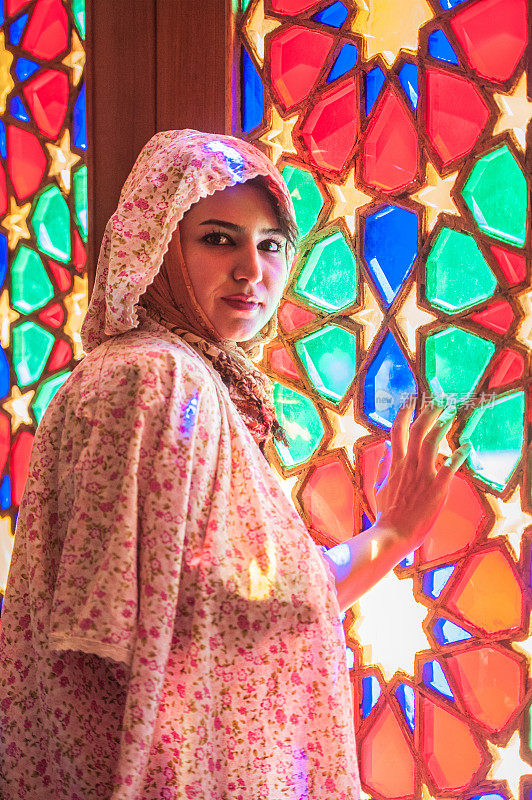 美丽的年轻伊朗妇女在传统穆斯林服装对着彩色彩色玻璃窗摆姿势，伊朗