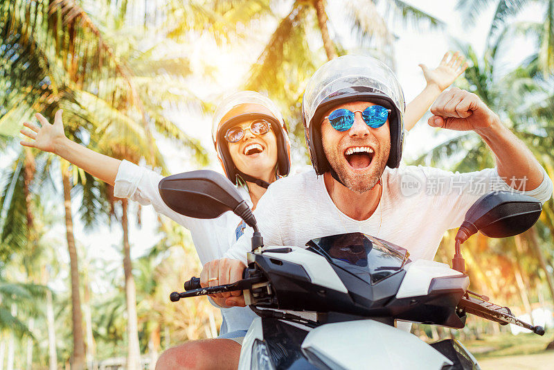 快乐微笑的夫妇旅行者骑摩托车在安全头盔在棕榈树下热带度假