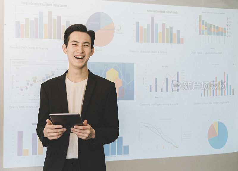 一名亚洲华裔年轻白领站在投影仪屏幕前，手里拿着一个数字平板电脑，笑着播放所有的预测和投影图