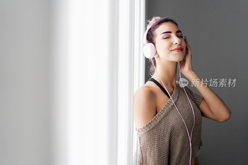 美丽的年轻女子在窗前戴着耳机欣赏音乐