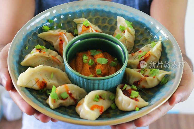 一个面目全非的人拿着一碗蒸饺(南亚饺子)，上面淋着辣椒油，点缀着葱花和香菜，周围是用橘子辣酱蘸酱的小模子，居高临下