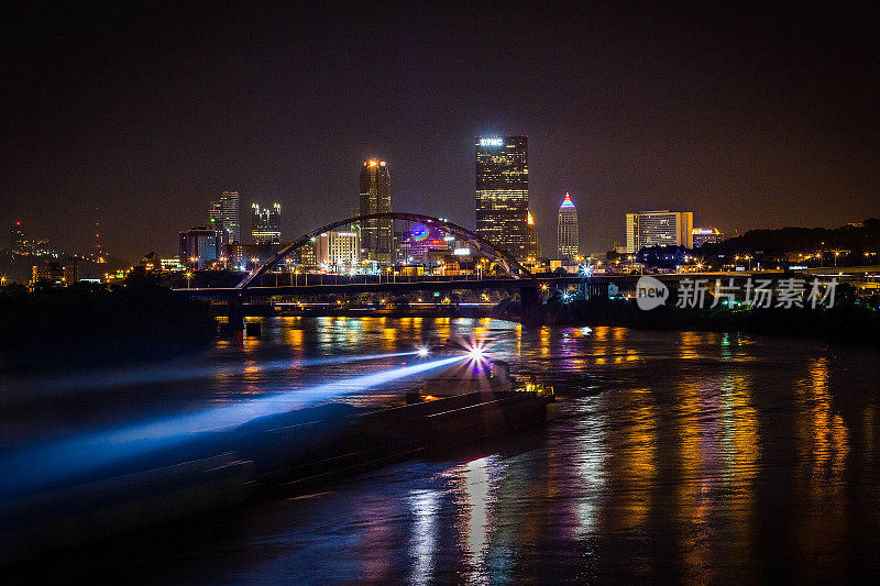 一艘驳船在夜晚穿过宾夕法尼亚州匹兹堡市时闪耀着它的聚光灯——在俄亥俄河后面，匹兹堡的天际线被明亮地照亮。