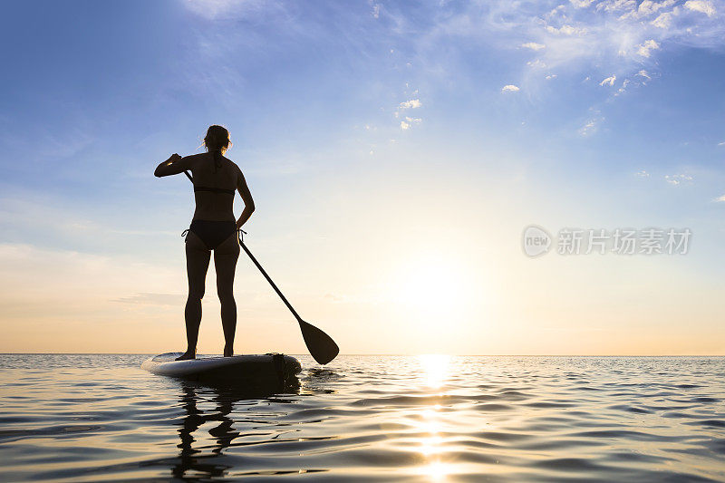 女孩站起来，桨板(sup)在平静的海面上，日落