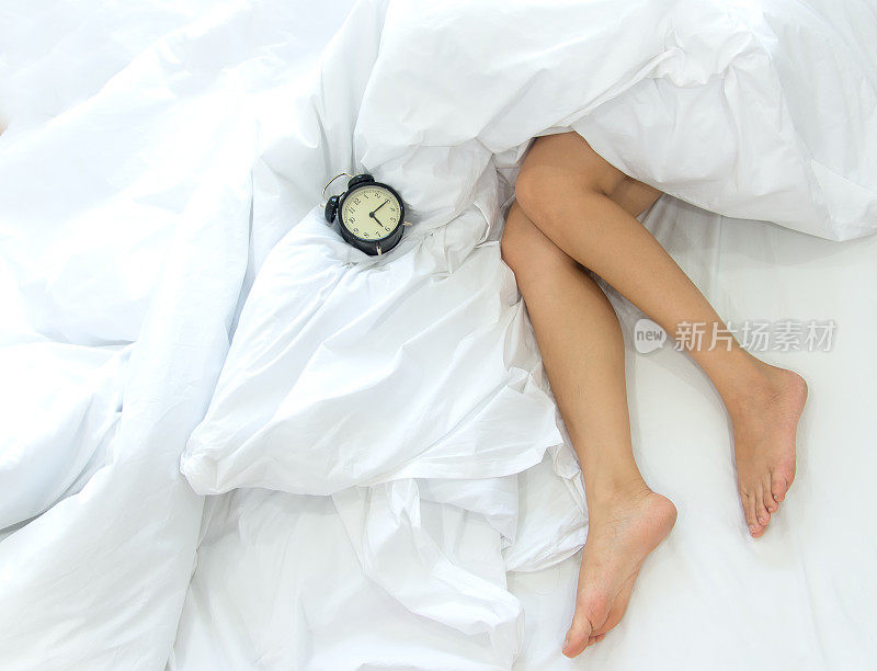 女性的腿和闹钟在床上的特写。懒惰的女人懒洋洋地躺在卧室里放松