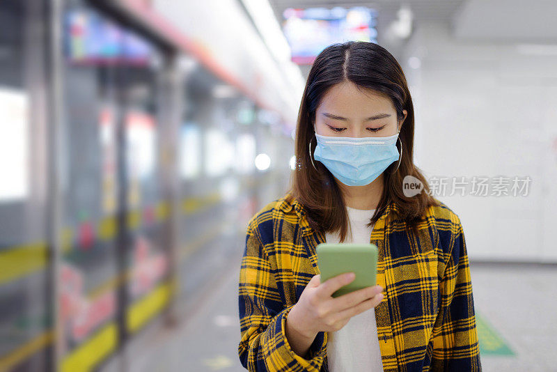 年轻的亚洲华人女子戴着口罩，在地铁站使用智能手机。