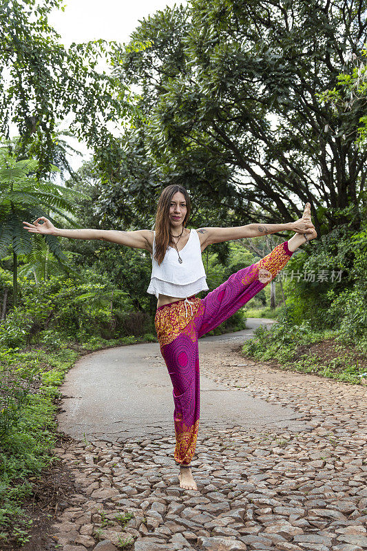 一名拉丁女子穿着色彩鲜艳的裤子，在绿树成荫的乡村公路上做瑜伽。