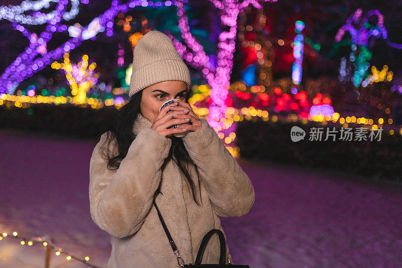 快乐的年轻女子一边喝咖啡一边享受户外的圣诞彩灯