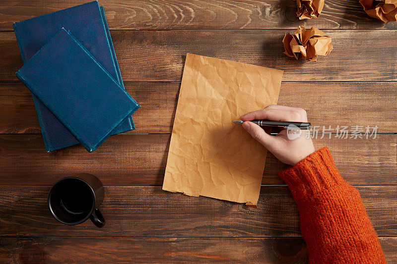 在白纸上写字。木桌上放着一杯咖啡、一本书和皱巴巴的纸。