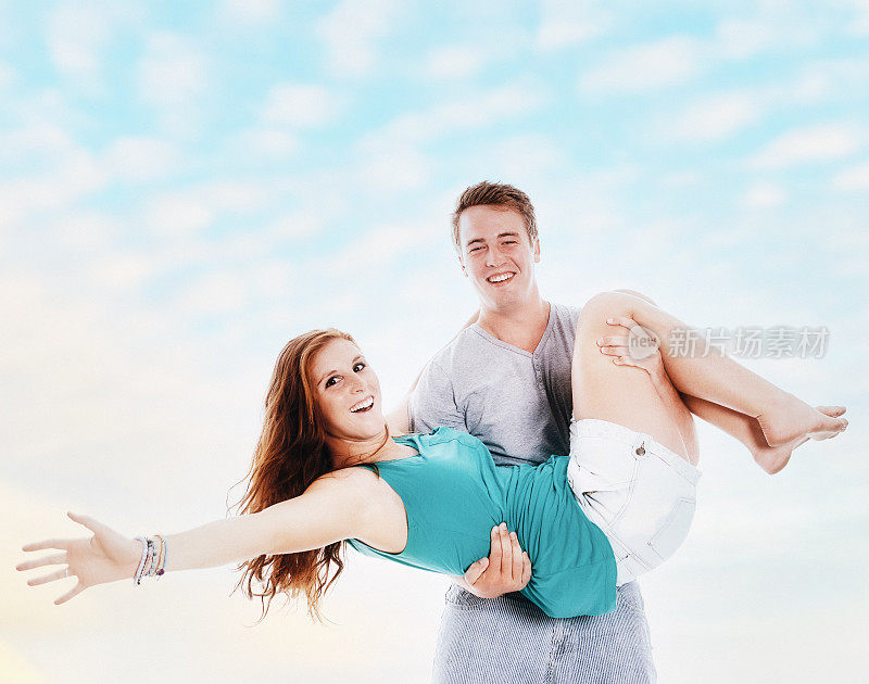 俏皮的年轻夫妇:笑着笑着，男人抱着女人在他的怀里，对着一个美丽的夏日天空与复制空间