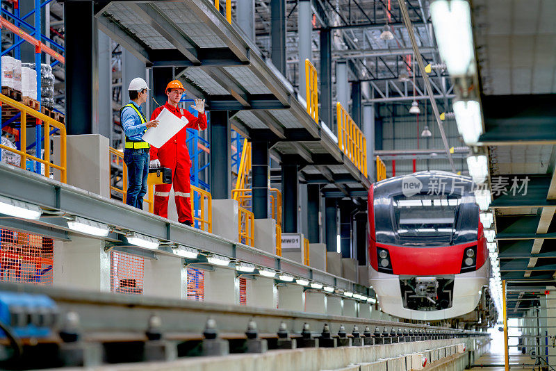 工程师手持图纸和技术工人在工厂车间电气或空轨列车的铁轨旁讨论。