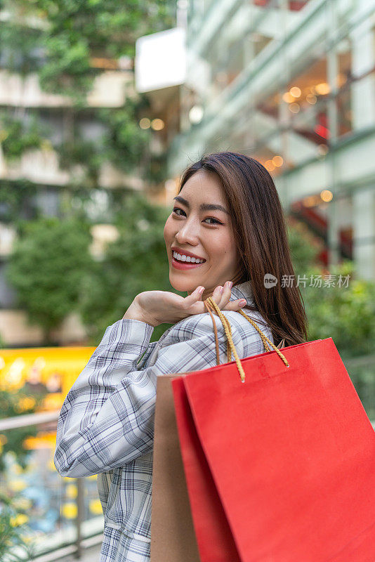 在一次成功的购物狂欢后，年轻的亚洲女商人在购物中心愉快地摆着购物袋，陶醉于购物疗法。