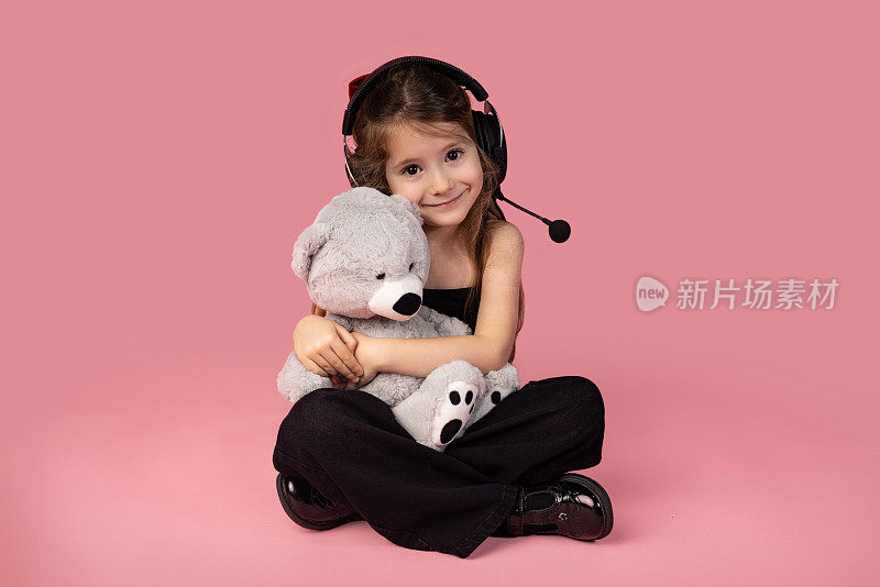 超级可爱的白人小女孩戴着耳机，看着镜头微笑着听音乐，拿着泰迪熊，激励孩子们去爱的概念。