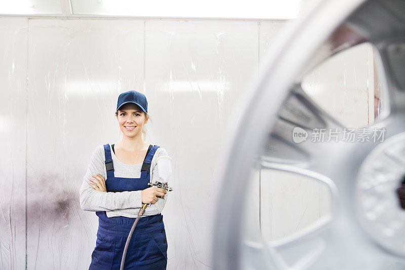 女汽车油漆工，在汽车车身车间油漆车辆。年轻女子穿着工作服，拿着喷枪，在轮辋上喷漆。