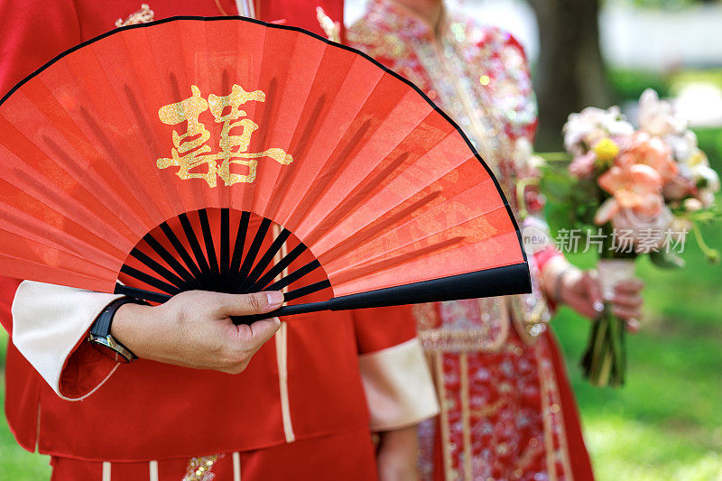 一对年轻的中国夫妇身着传统的结婚礼服，手持一把饰有“双喜”标志的红手扇子