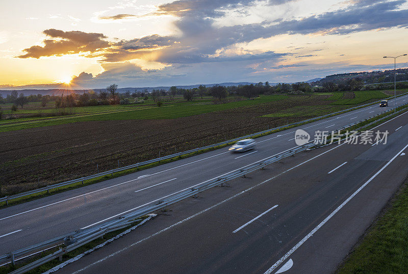 两辆汽车在公路上高速行驶，穿过乡村风景。高速公路高速行驶。高速公路上超速行驶的景象。背景是美丽的日落。