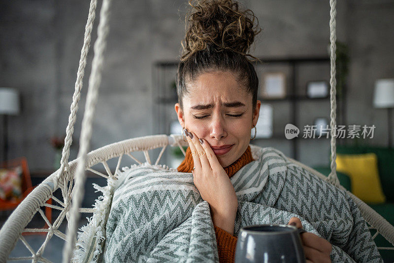 年轻女子喝着热茶或咖啡，感到牙痛，坐在家里客厅的秋千上。