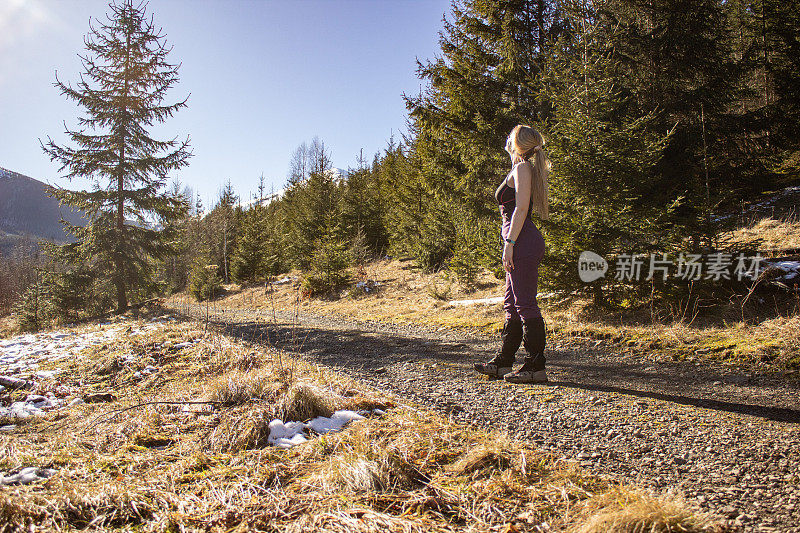 年轻的女徒步旅行者侧着身子站着，在森林的背景下，她的头转向太阳