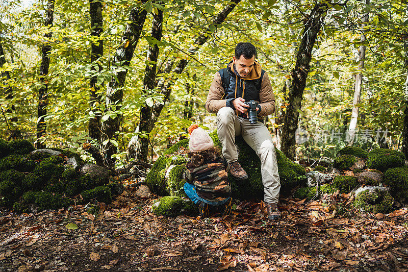 背着背包的摄影师父亲和戴着帽子的时尚儿子走完小路，坐在森林里的一块石头上