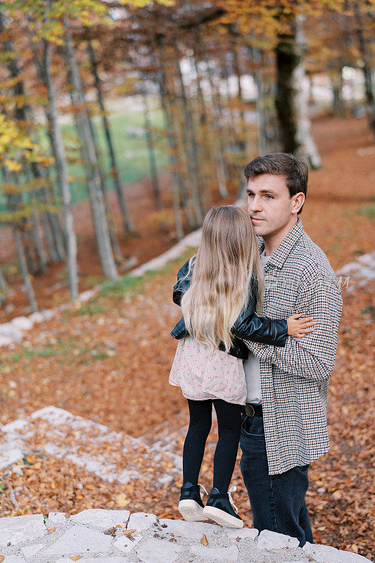 爸爸抱着一个站在秋林小山上的石径上的小女孩，望着远方