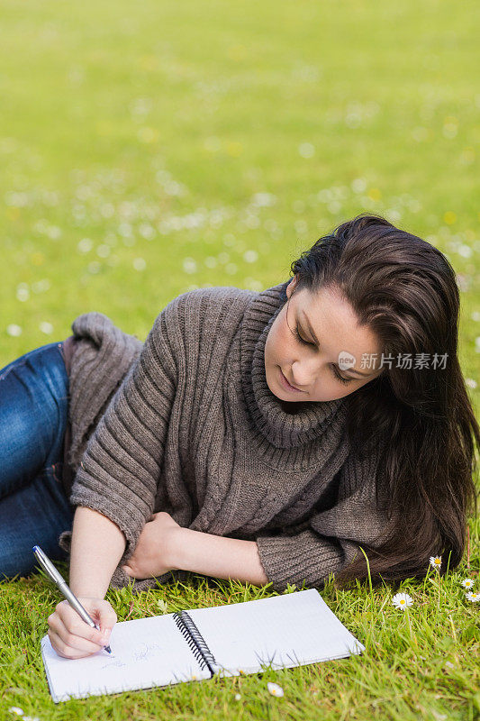 体贴漂亮的棕发女孩躺在草地上写作