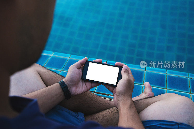 特写一名男子手持手机与空白拷贝空间屏幕上，与腿浸在游泳池在夏季度假。手机屏幕上的剪切路径