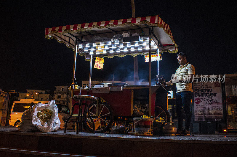 中东，土耳其，伊斯坦布尔:城市的夜生活和街头小吃，晚上在伊斯坦布尔的街道上有一个红白条纹的小摊，一个烤煮玉米小贩