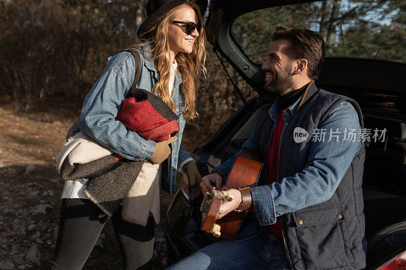 在他们的公路旅行冒险中，一个男人在汽车后备箱旁给他的未婚妻弹吉他