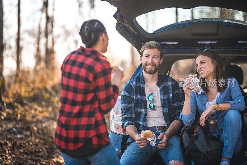 在森林里，朋友们在汽车后备箱边聊天，吃三明治