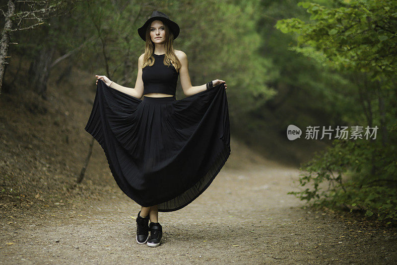 一个金发年轻女子的肖像戴着一顶帽子穿着一件黑色的衣服在树林里。有魅力的女孩在散步。现代女性的时尚
