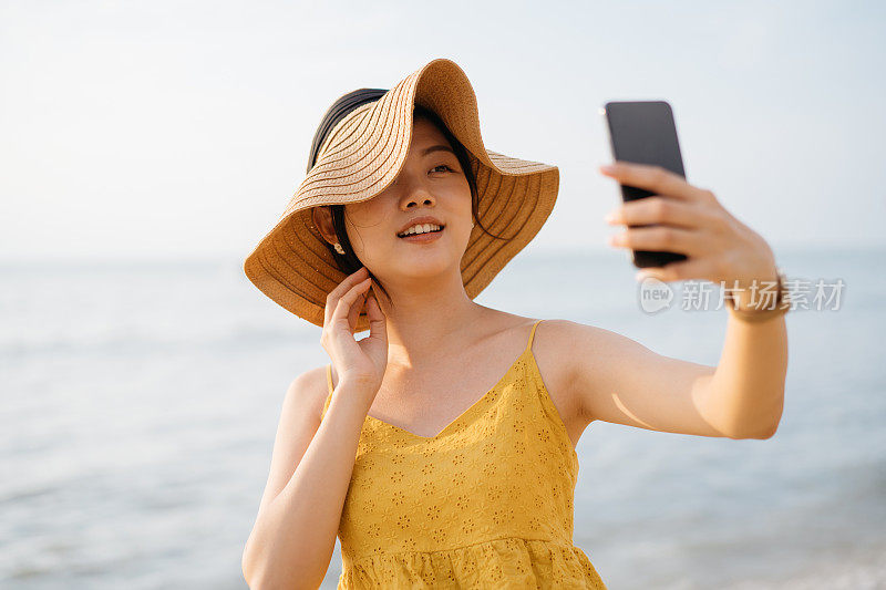 年轻的亚洲女游客戴着太阳帽用智能手机在海滩自拍