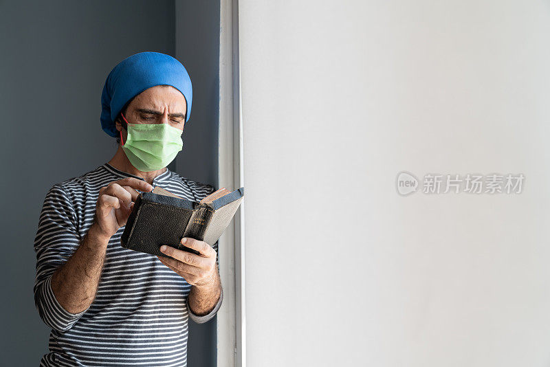 成年男子戴着防护面具站在窗边阅读圣经