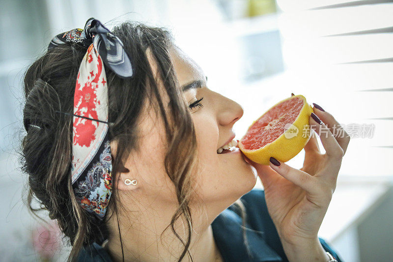 一个年轻的女人正拿着一个葡萄柚坐在她的厨房里，享受着一个非工作的日子-一个快乐的年轻女孩拿着一个葡萄柚-一个全方位的美容助推器
