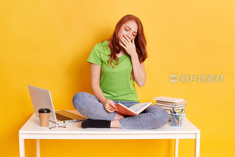 可爱的红头发疲倦困倦的女人坐在她的桌子上，旁边放着书和电脑，闭着眼睛，打哈欠时捂住嘴，靠着黄色的墙花很长时间学习。
