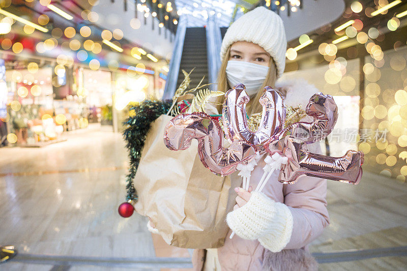 年轻女子戴着医用口罩在商场为圣诞节购物。手持数字2021的金色气球的女性。新冠肺炎疫情下的圣诞假期。有选择性的重点