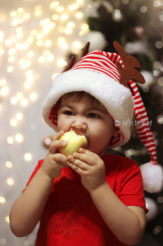 戴着圣诞帽的小男孩在圣诞彩灯的背景下咬苹果