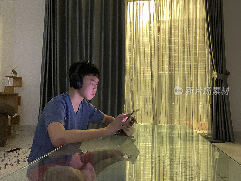 年轻的亚洲男孩戴着耳机在起居室用数字平板电脑玩视频游戏