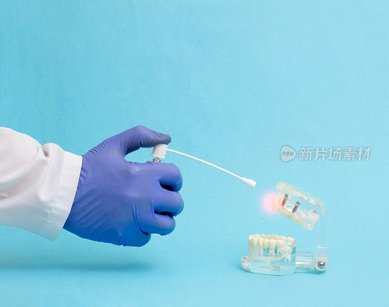 蓝色背景上的牙医医生的手拿着麻醉喷雾剂，靠近一个有牙齿的下颌模型。局部麻醉概念在牙科，复制空间的文本，牙周病