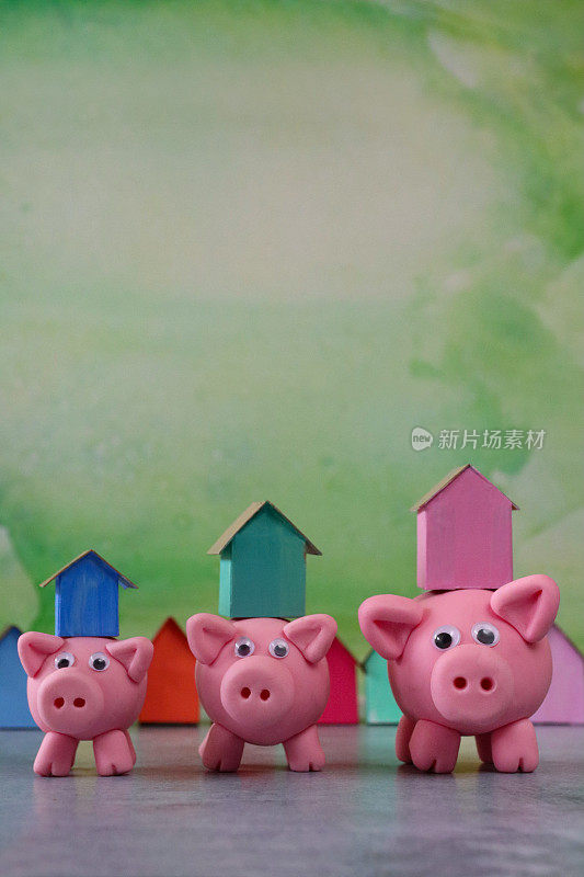 三只模型猪的形象与模型纸板房子平衡在小猪存钱罐的背部，排彩色的海滩小屋，重点在前景，斑白的绿色背景，家庭财务，房地产和假日储蓄概念，复制空间